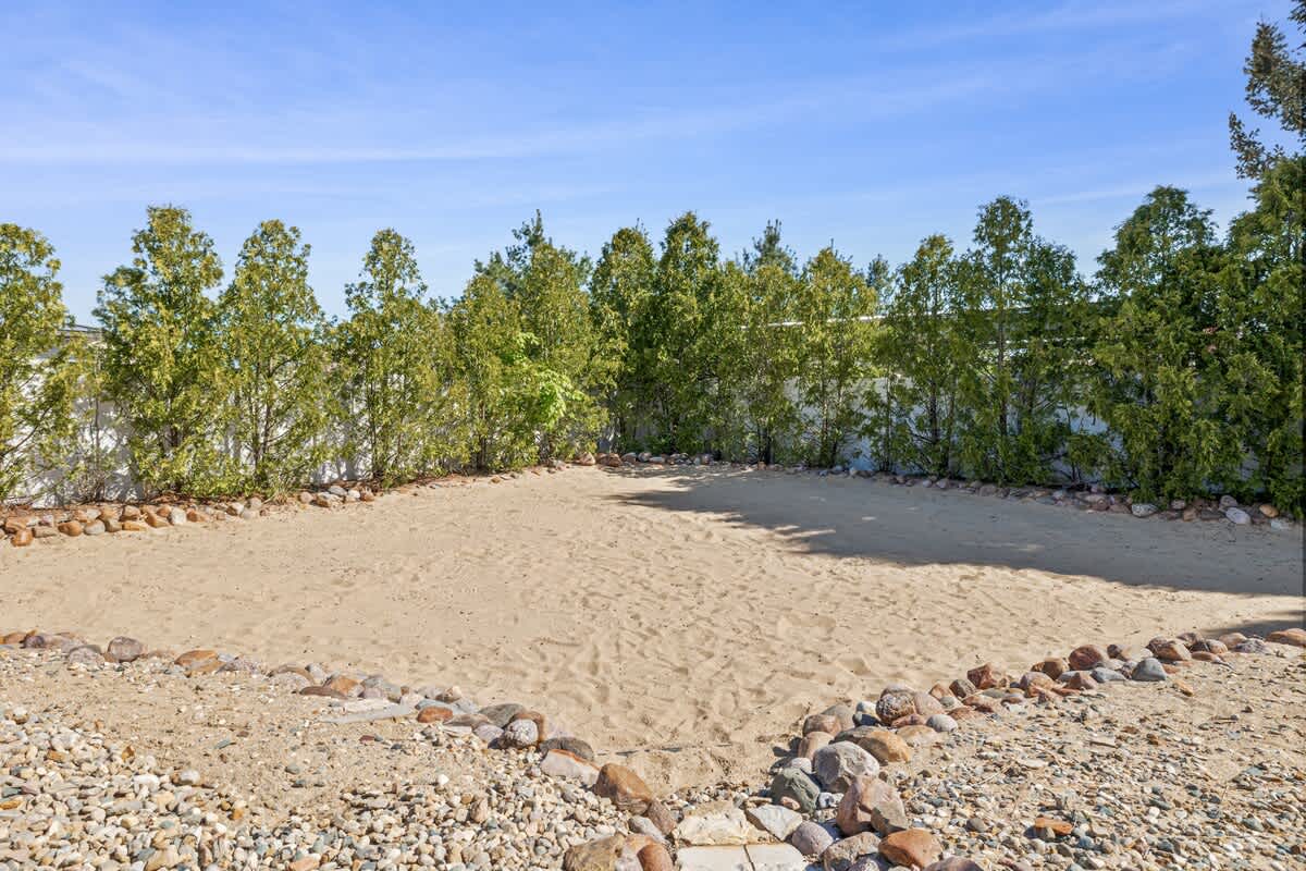 A large, rectangular sand pit area in a backyard, bordered by a decorative rock perimeter. Tall green hedges line the back, providing privacy and a natural backdrop for outdoor activities.
