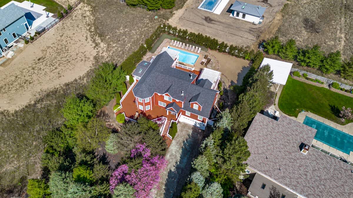An aerial view showcasing the red-sided house with a dark roof, surrounded by lush green trees and landscaping. A swimming pool is visible in the backyard, and the property is situated within a residential area with other homes and undeveloped land.