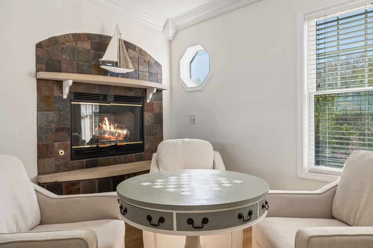 A cozy corner of the living room featuring a stone-surround fireplace with a mantel decorated with a sailboat model, flanked by two upholstered armchairs. A round game table with a checkerboard pattern is centered between the chairs.