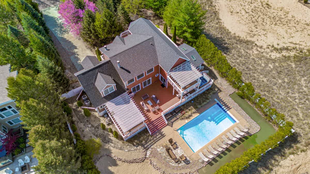 An aerial view showcasing a large, multi-level house with a dark roof, a spacious deck with a pergola, and a bright blue swimming pool. The property is surrounded by trees and sandy areas.
