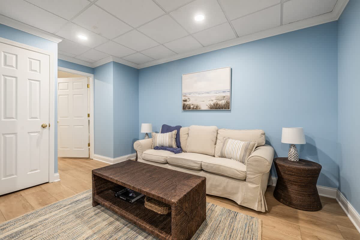 Inviting living area with light blue walls, light wood floors, and a beige sofa adorned with pillows. A dark woven coffee table sits on a striped rug, and a door leads to another room.