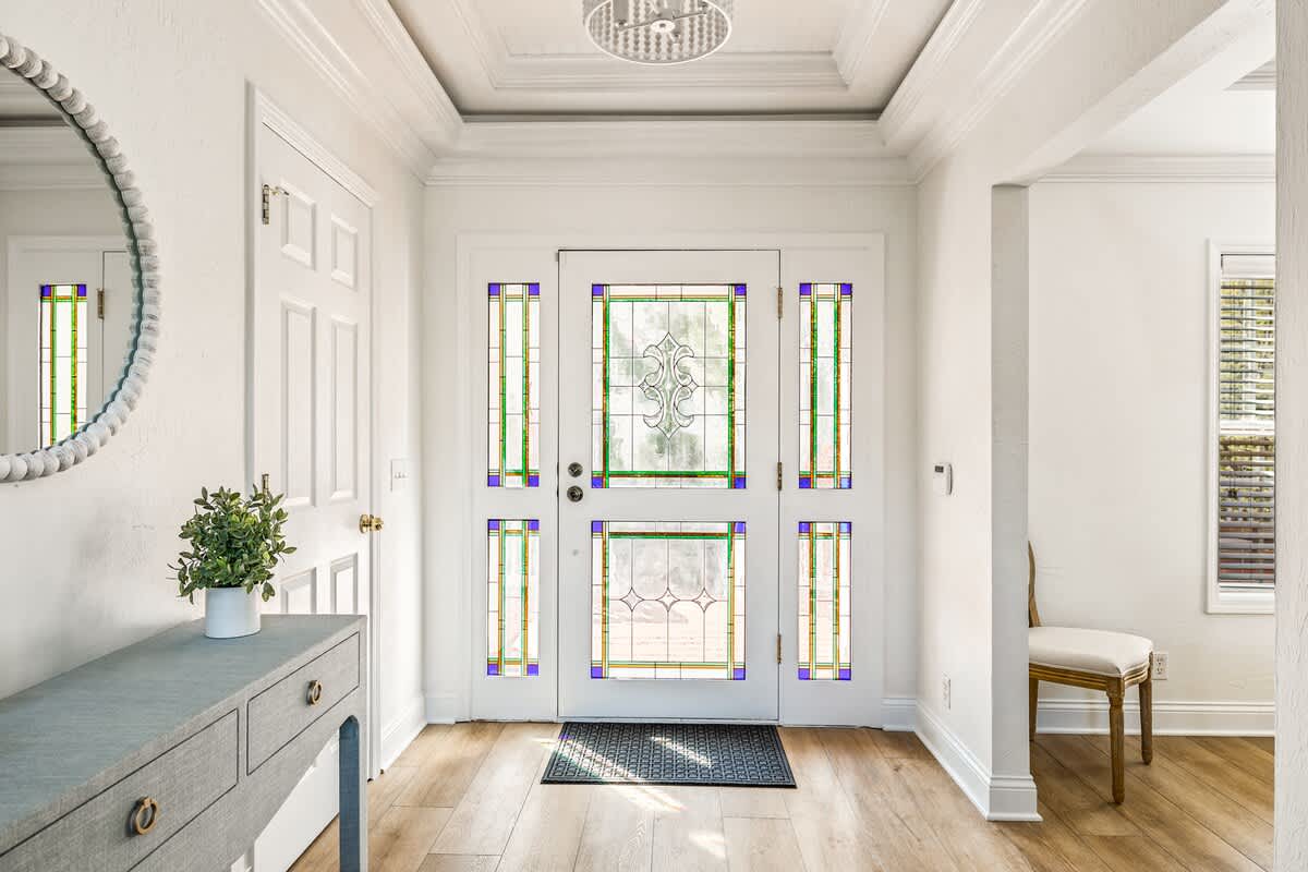 A welcoming foyer with light wood flooring and white walls, featuring a front door with intricate stained glass panels and a decorative round mirror above a grey console table.