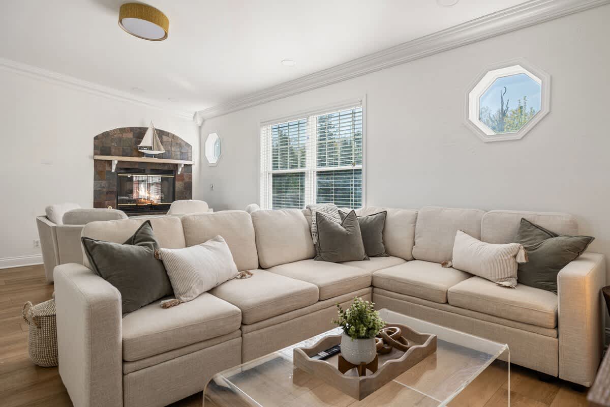 A spacious living room with light wood floors, featuring a large L-shaped sectional sofa in a neutral tone with olive green and white accent pillows. A clear acrylic coffee table sits in front, and a fireplace with stone surround is visible in the background.