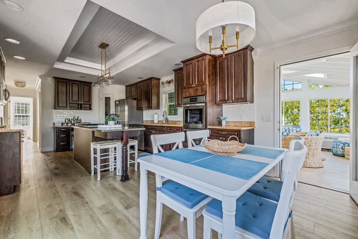 Open-plan kitchen and breakfast nook area with dark wood cabinets and a white dining set, offering a glimpse into an adjacent bright sunroom or lounge area through an open doorway.
