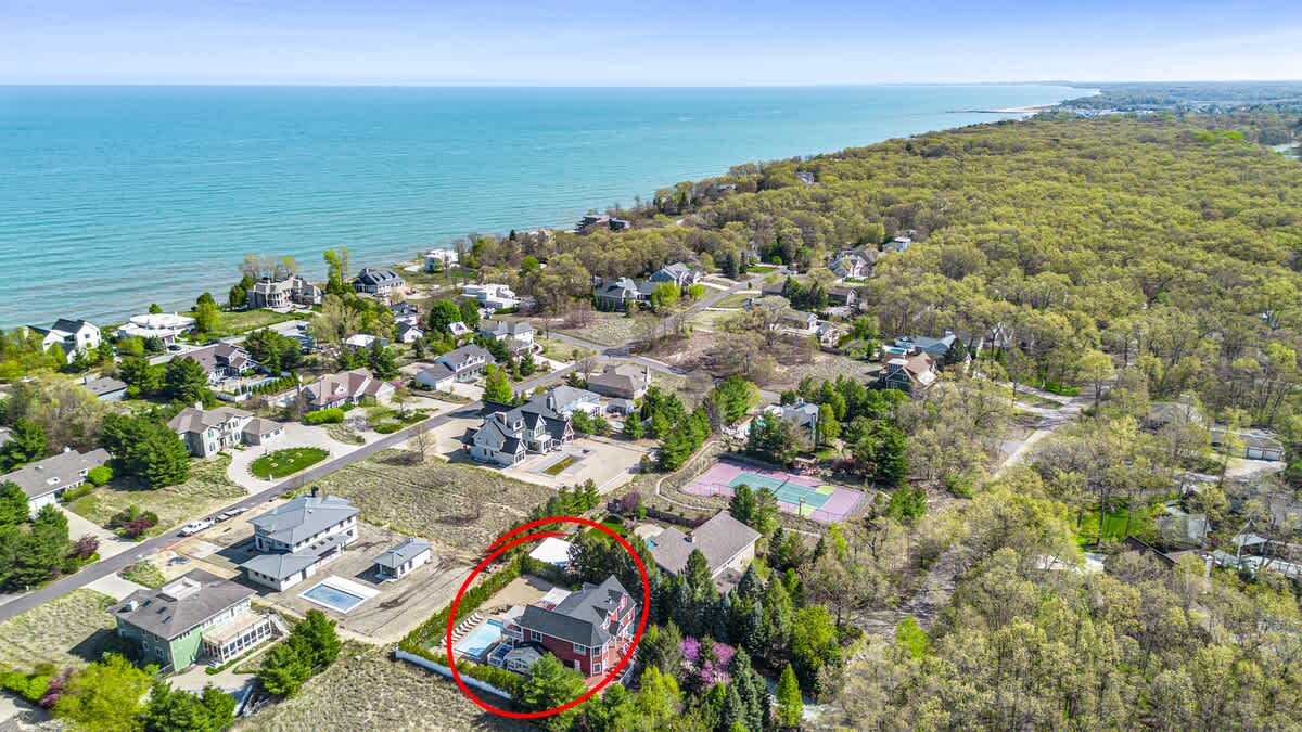 A broad aerial perspective of the property, circled in red, illustrating its close proximity to a vast blue lake or ocean and a long coastline. The surrounding area features numerous houses, dense forests, and open land under a clear sky.
