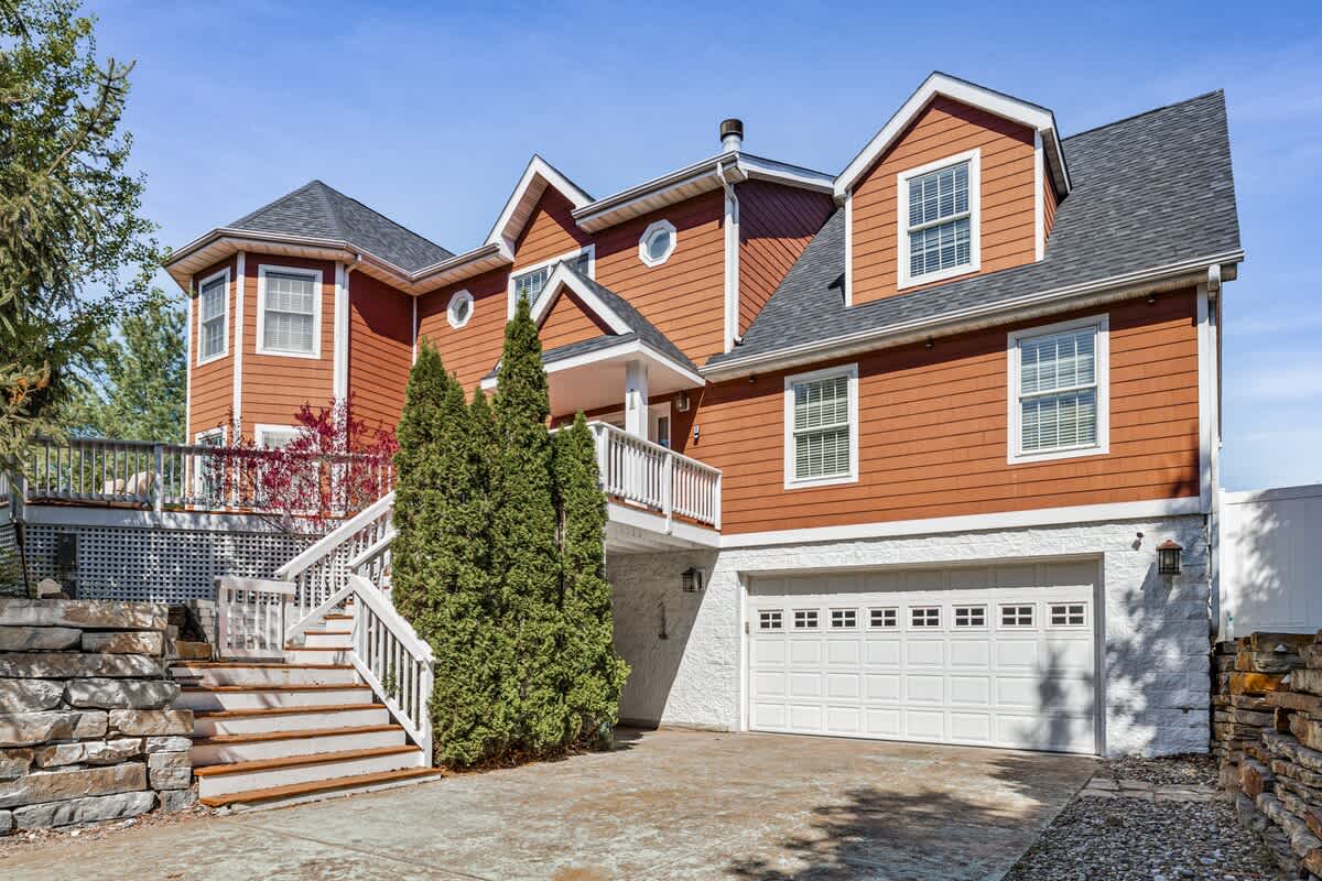 The front exterior of a multi-story house with reddish-brown siding and a dark roof, featuring a white garage door, a stone retaining wall, and a prominent white staircase leading to an upper deck.