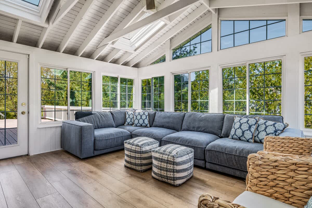 Bright sunroom living area with a large gray sectional sofa and two patterned ottomans, featuring numerous windows with grid patterns, skylights, and a vaulted white wood-paneled ceiling.