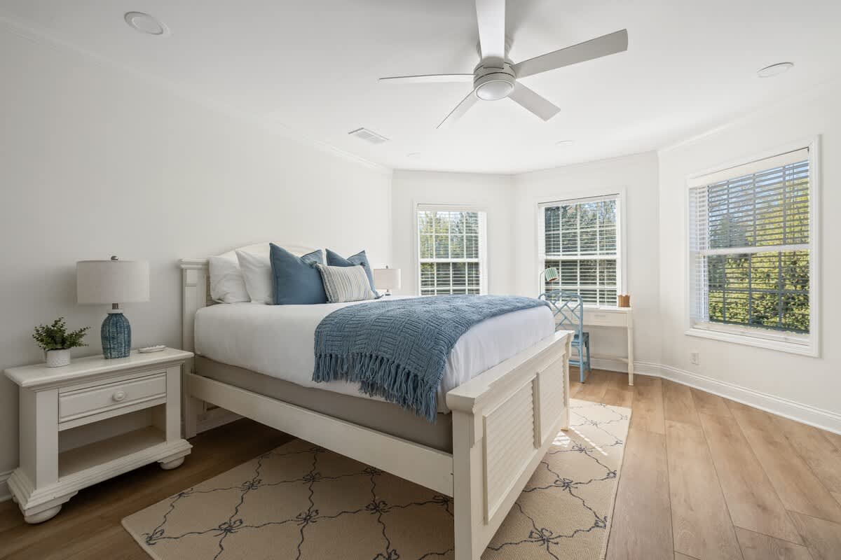 Serene bedroom featuring a white bed frame with blue and white bedding, light wood floors, and multiple windows with blinds. A white nightstand with a lamp is visible on the left.