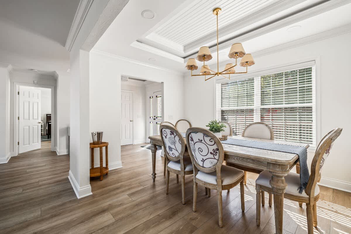Spacious dining room with a light wood dining table and upholstered chairs, a brass chandelier, and a view towards the front entrance and a hallway, showcasing light wood flooring and white trim.