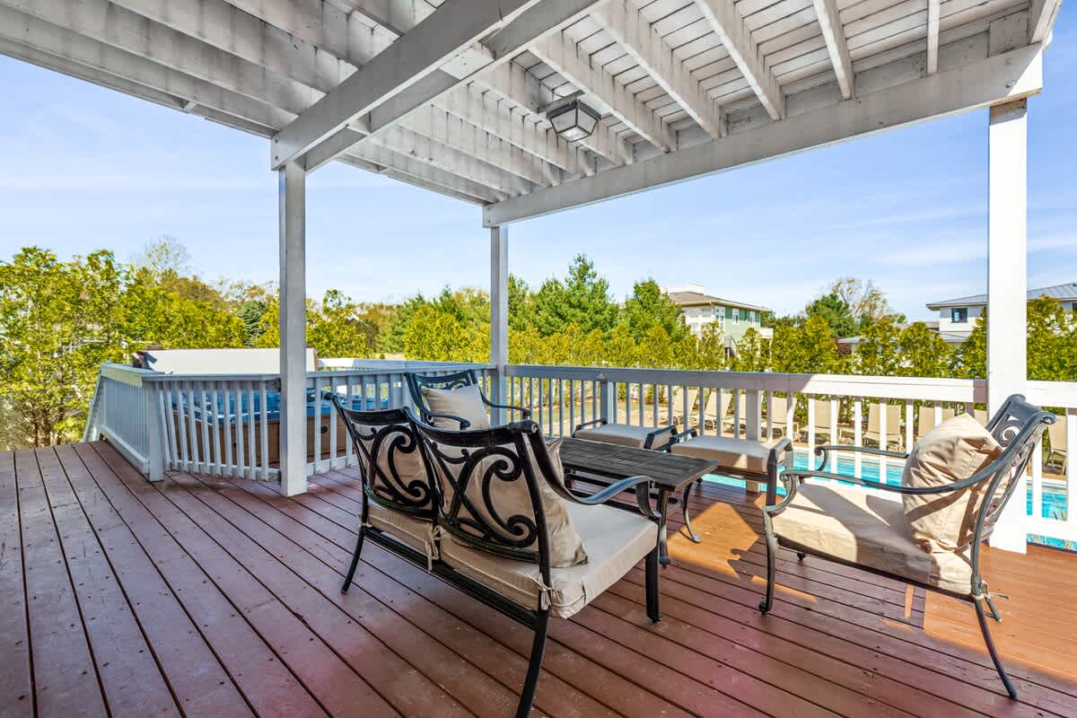 Covered upper-level balcony with a red-stained wooden floor, white railings, and a small seating area, offering a view of the pool and a hot tub on a lower deck.