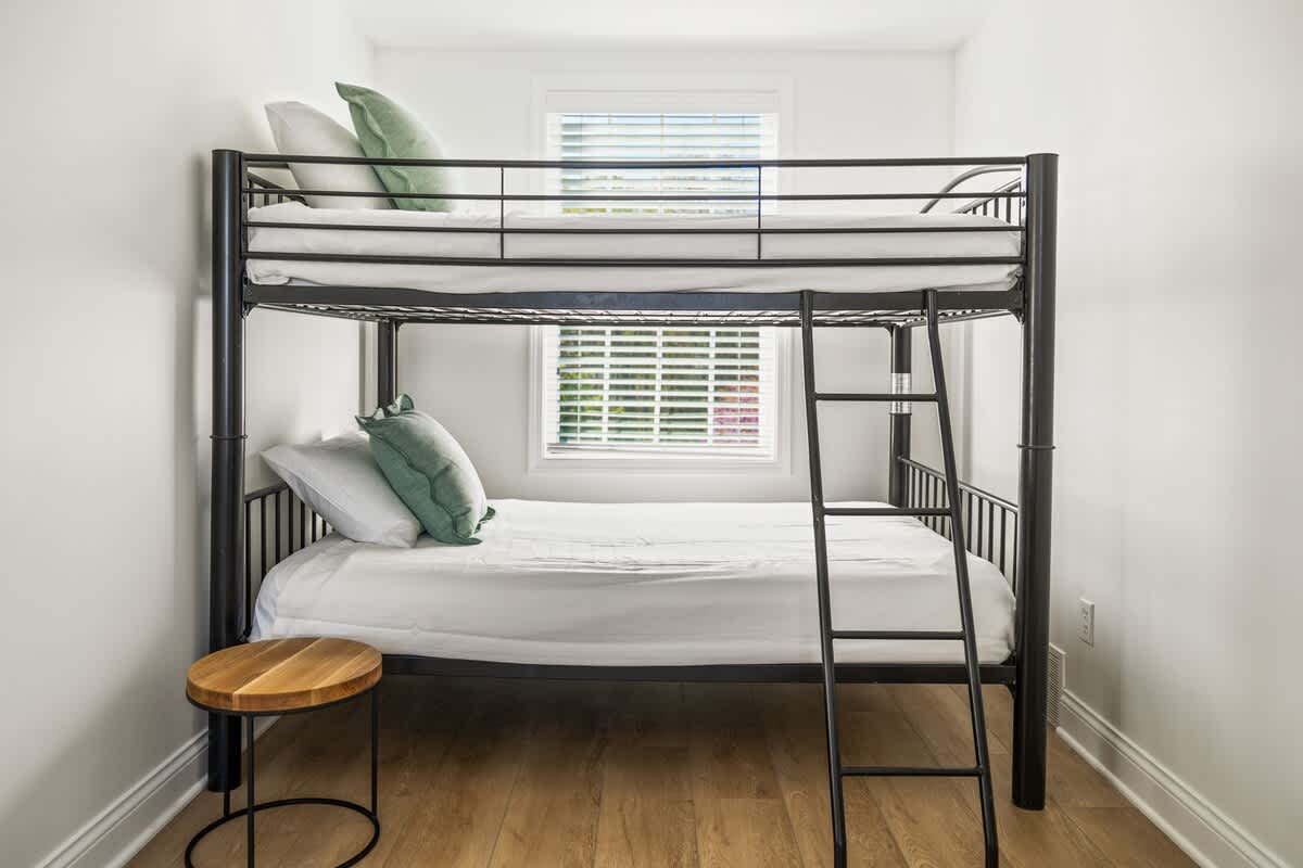 A close-up view of a black metal bunk bed with white bedding and green accent pillows, positioned against a white wall. A small round wooden side table sits next to the lower bunk.