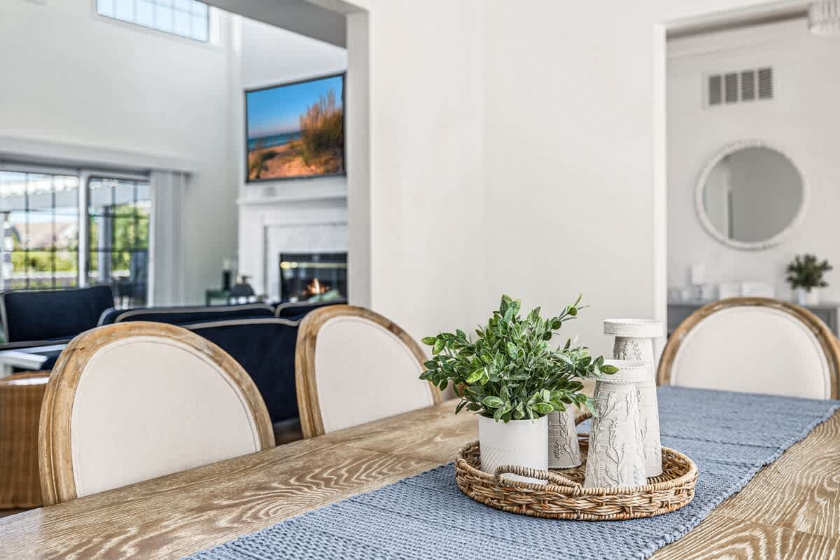 Close-up of a rustic wooden dining table adorned with a blue runner and a woven tray holding a potted plant and decorative candles, with the living room and a wall-mounted TV visible in the background.