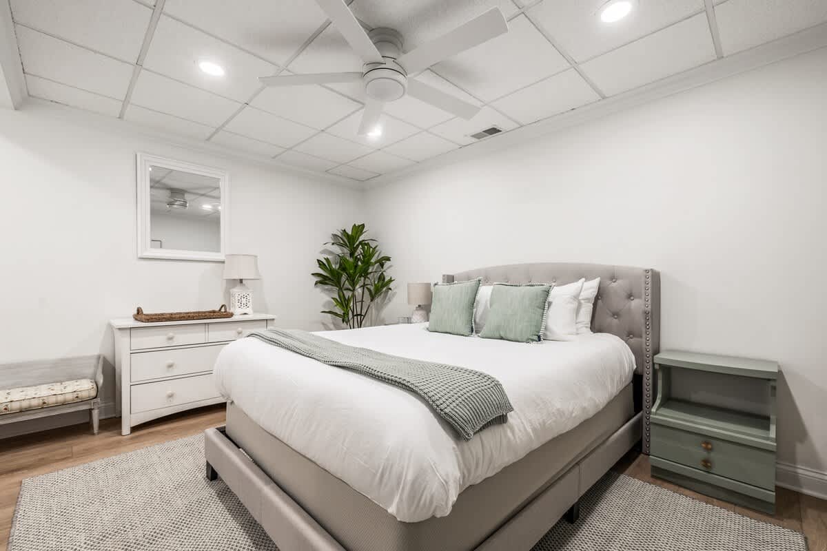 A bright bedroom featuring a light gray upholstered bed with a white duvet, a white dresser with a mirror above it, and a large plant in the corner. A ceiling fan and recessed lighting illuminate the room.