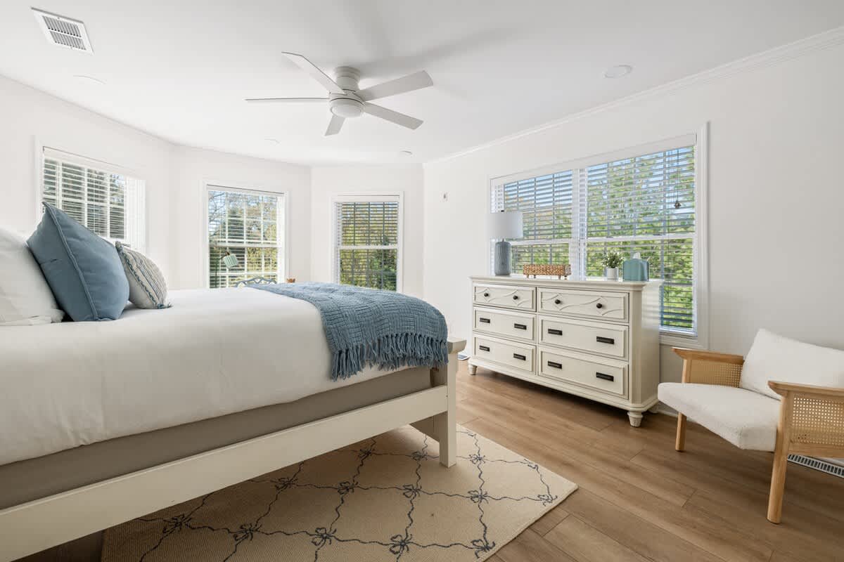Bright bedroom with a white bed, light wood floors, and a white dresser with a lamp. Multiple windows with blinds overlook greenery, and a rattan armchair adds a cozy touch.