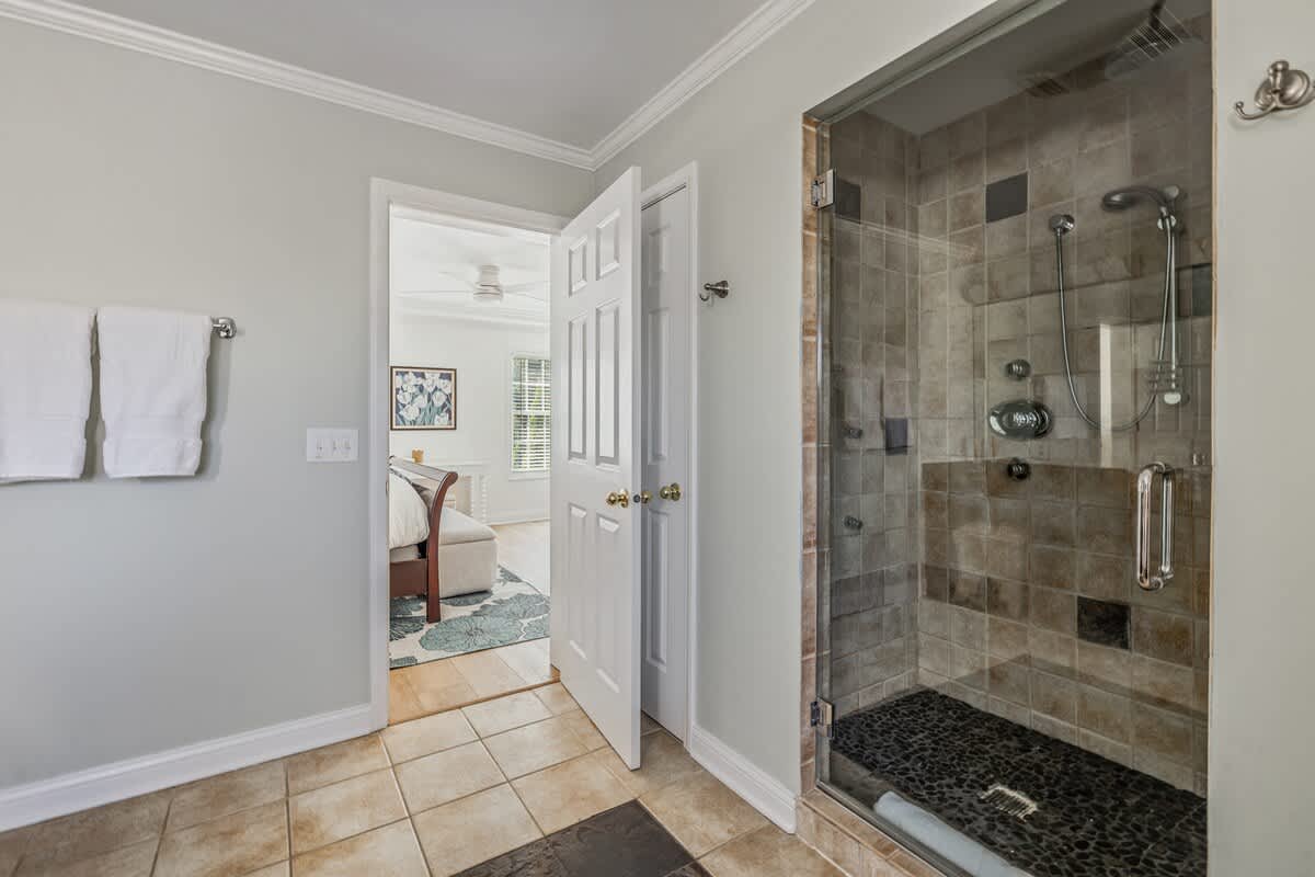 A modern bathroom featuring a glass-enclosed walk-in shower with tiled walls and a dark pebble floor, adjacent to a doorway leading to a bedroom. White towels hang on a rack against light grey walls.
