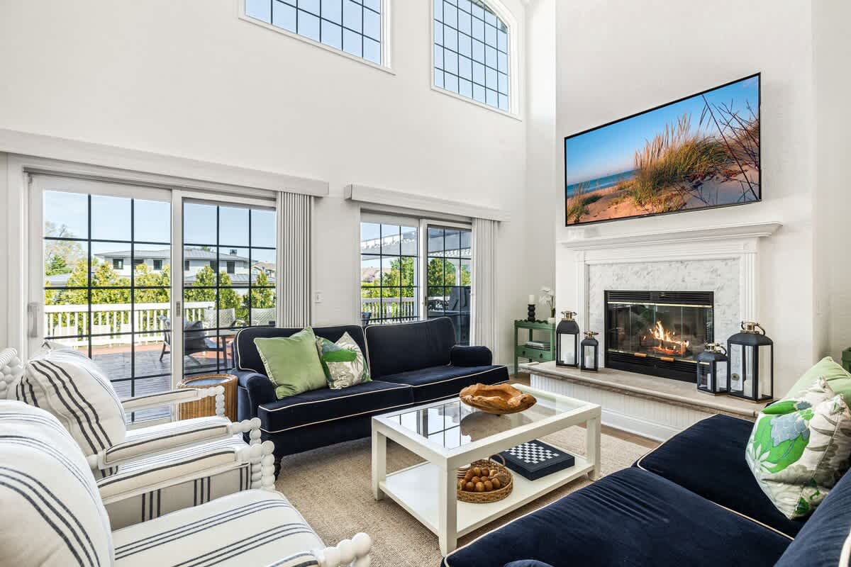 A spacious living room with high ceilings, white walls, and light flooring, centered around a marble fireplace with a TV mounted above. Two dark blue sofas and striped armchairs face a glass coffee table.