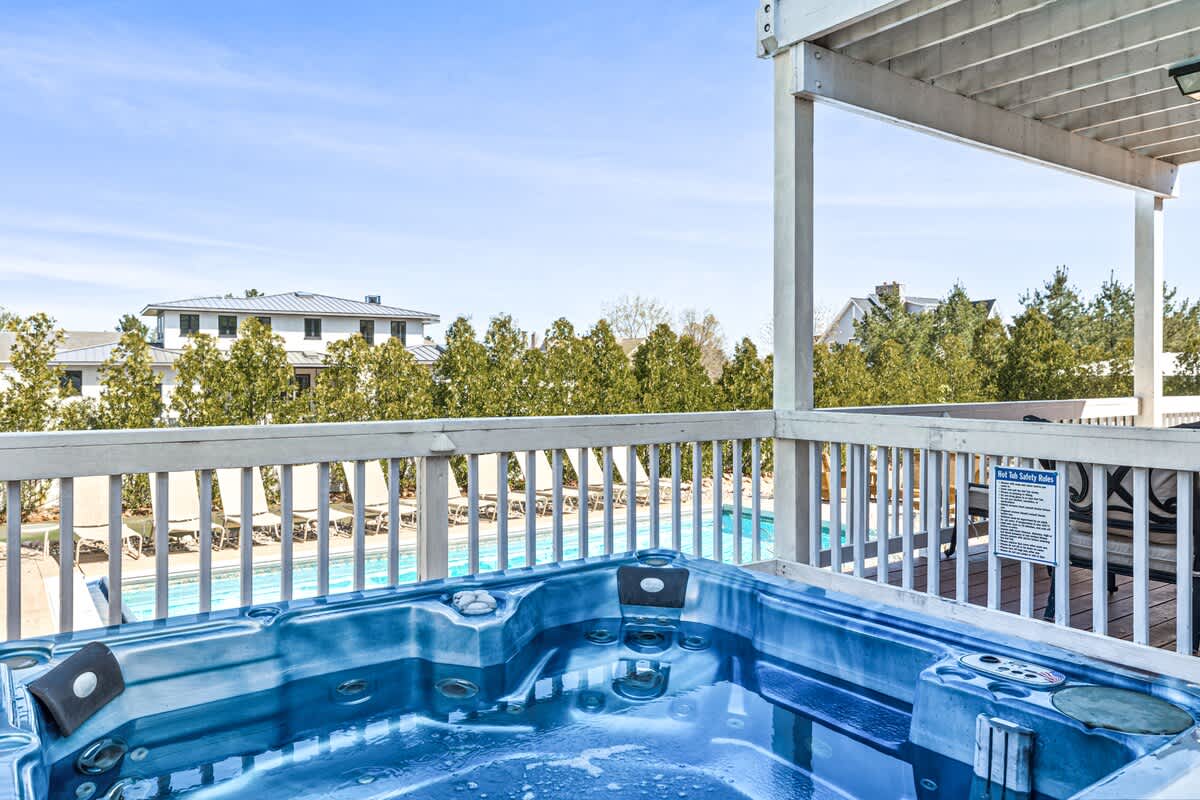 A blue hot tub is positioned on a wooden deck with white railings, offering a view of the swimming pool and surrounding greenery under a clear sky.