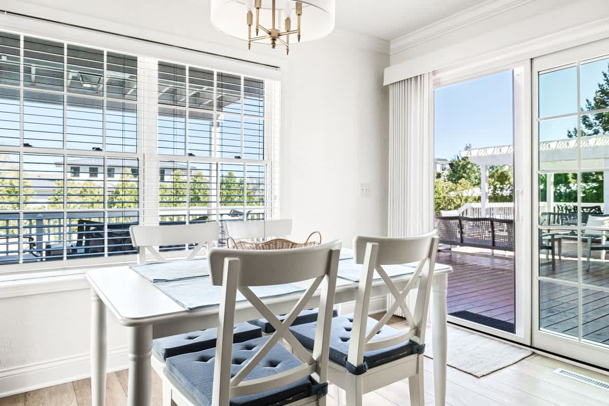 Sunny dining area featuring a white rectangular table with blue placemats and white chairs, positioned between large windows with white blinds and a sliding glass door opening to an outdoor deck.
