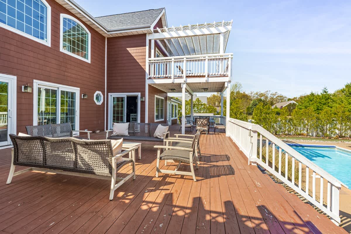 The back of a multi-story house with reddish-brown siding, featuring a large wooden deck with white railings and multiple seating areas. A swimming pool is visible at the bottom right, and a second-story balcony with a white pergola overlooks the deck.