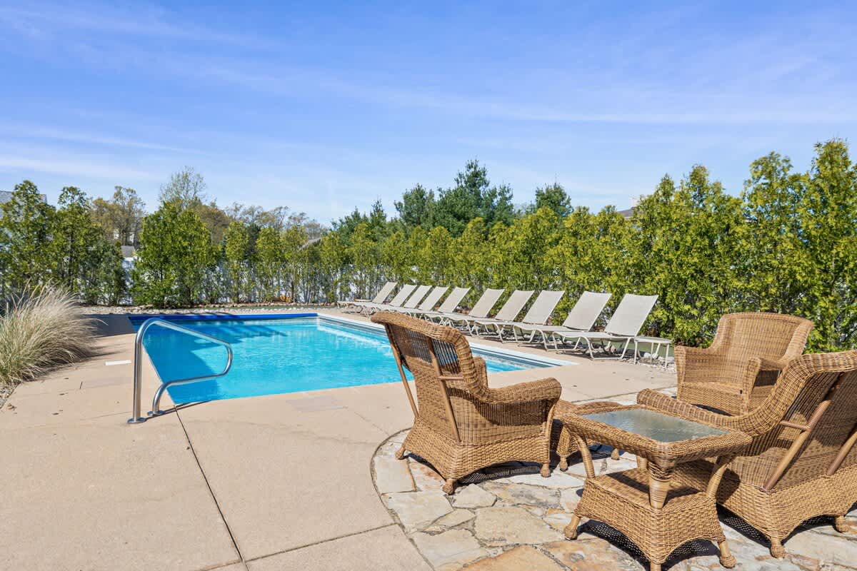 A spacious outdoor pool area featuring a rectangular swimming pool with a metal handrail, surrounded by a concrete and flagstone patio. A set of wicker armchairs and a matching table create a comfortable seating arrangement near the pool.