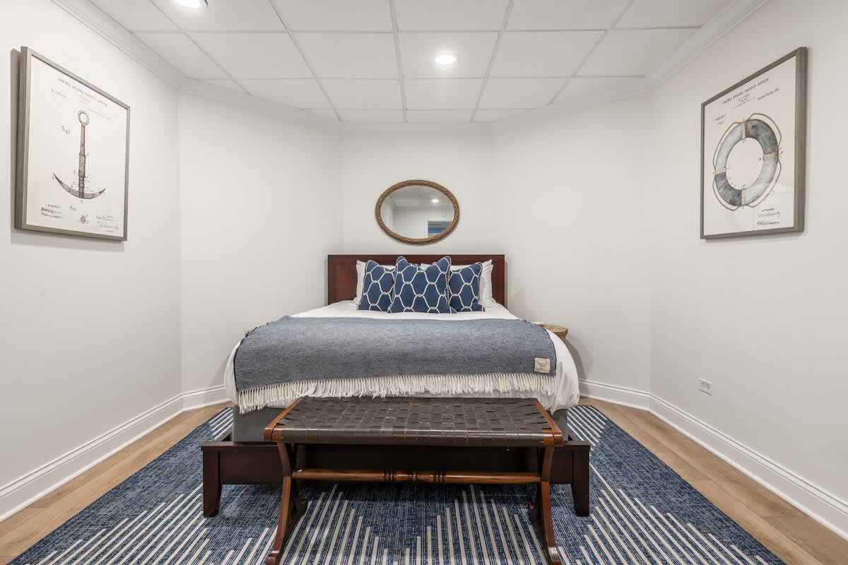 A cozy bedroom with white walls and a dark wooden bed frame, featuring a blue and white patterned rug and two framed nautical-themed artworks on the walls. A dark brown leather bench sits at the foot of the bed.