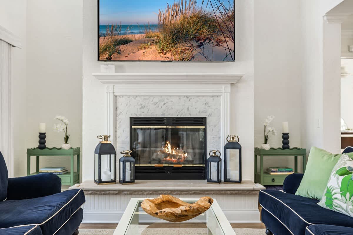 A detailed view of a white marble fireplace with a burning fire, flanked by two green side tables and decorative lanterns. A large TV is mounted above the mantel.