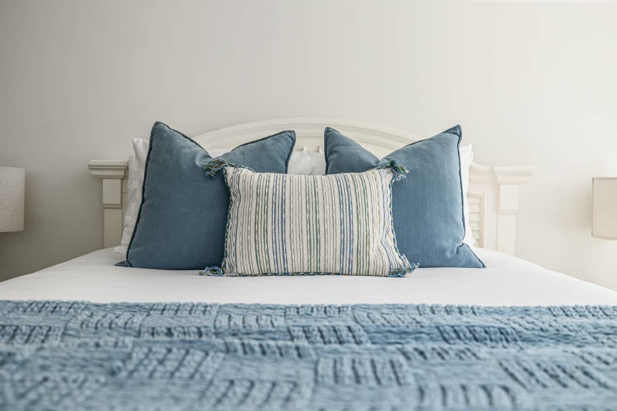 Close-up of a neatly made bed with a white headboard, featuring two large blue pillows and a striped rectangular pillow with tassels. A blue textured throw blanket covers the foot of the bed.