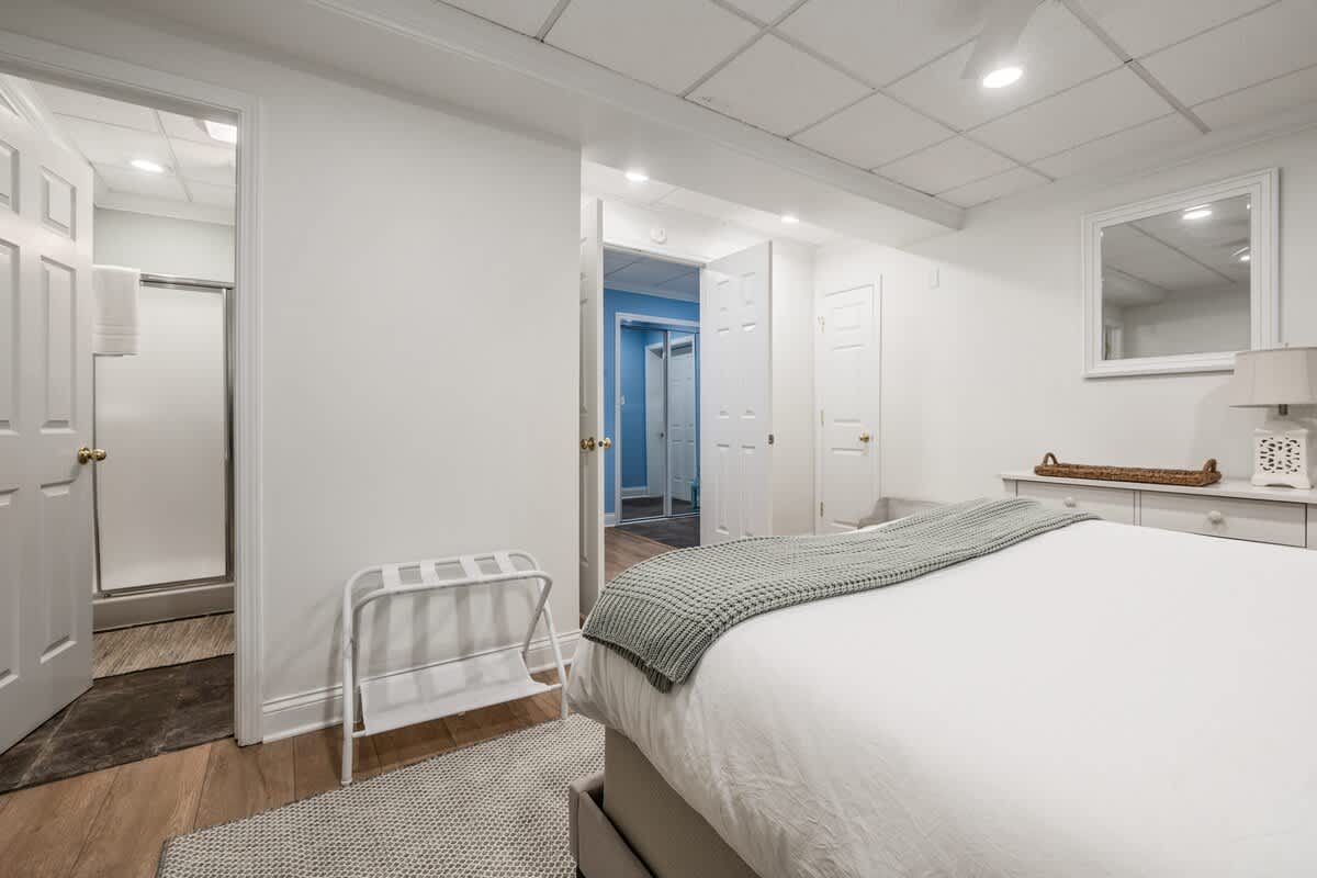 A view of a bedroom with a light gray upholstered bed and a white dresser, showing a doorway leading to an adjacent bathroom with a glass-enclosed shower. A luggage rack is placed near the doorway.
