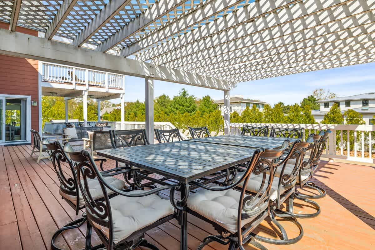 Large outdoor deck with a white wooden pergola providing partial shade over two dark metal dining tables with cushioned chairs, offering views of surrounding greenery and distant houses.