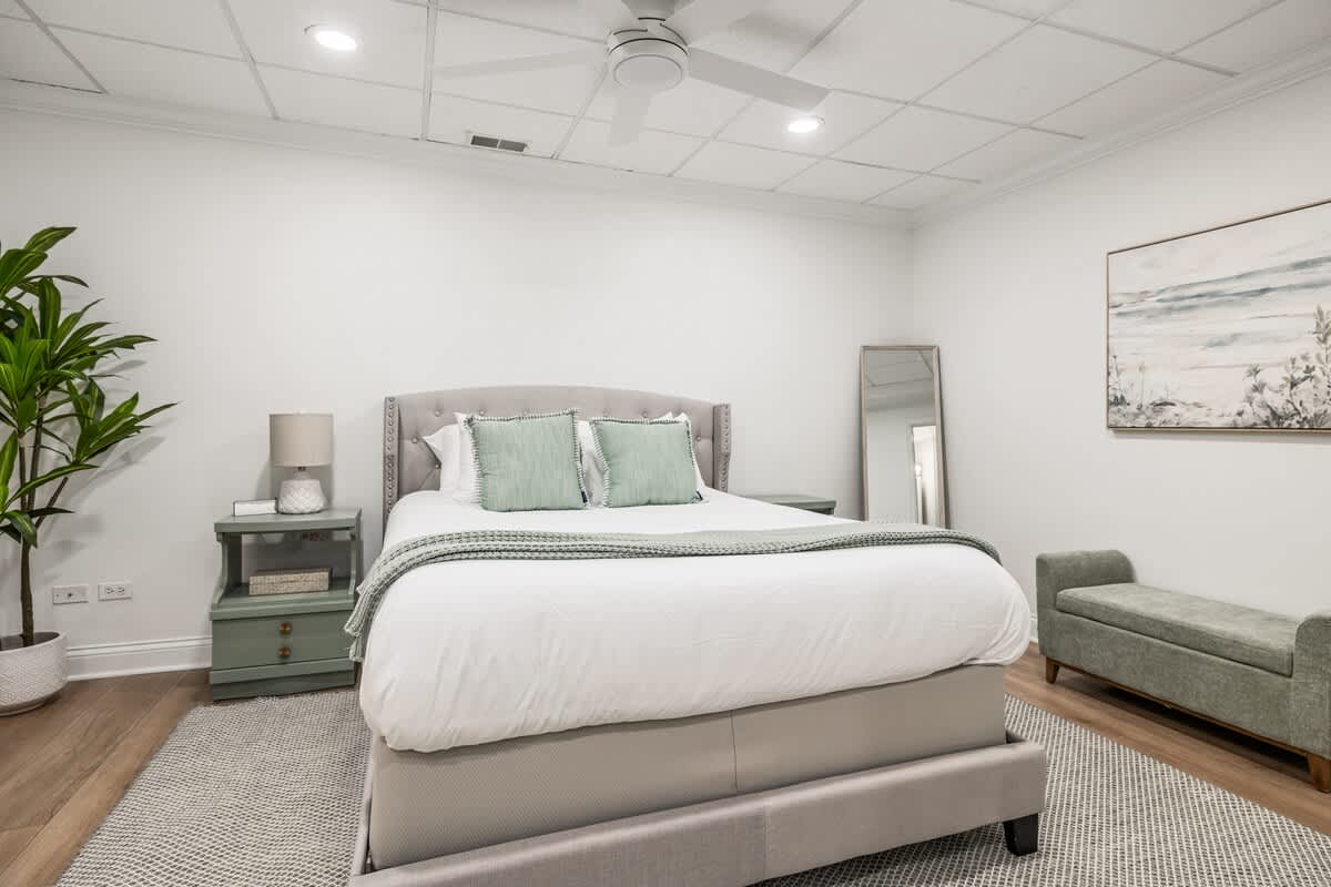 A spacious bedroom with a light gray upholstered bed, a textured gray area rug, and light green nightstands. A large plant and a full-length mirror are visible, creating a serene and comfortable atmosphere.