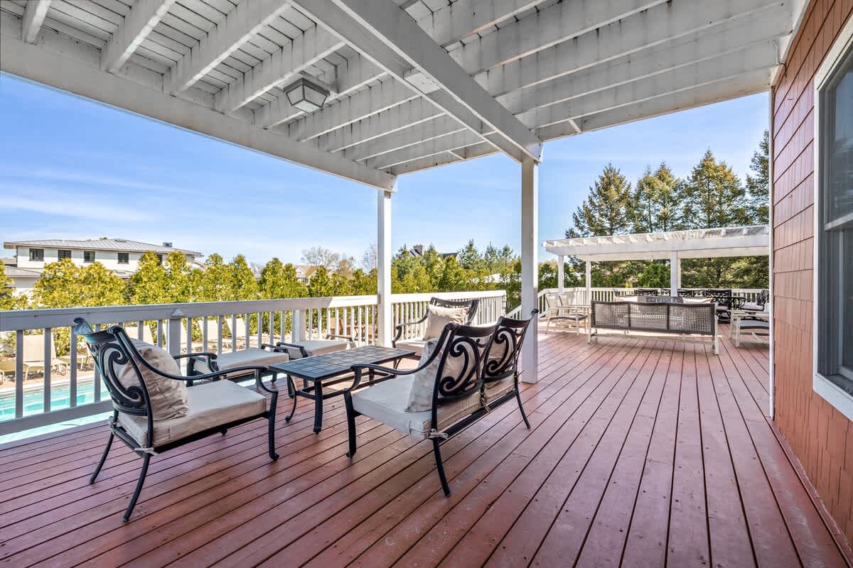 Covered upper-level balcony with a red-stained wooden floor and white railings, furnished with dark metal chairs and a small table, overlooking a pool and lush trees.