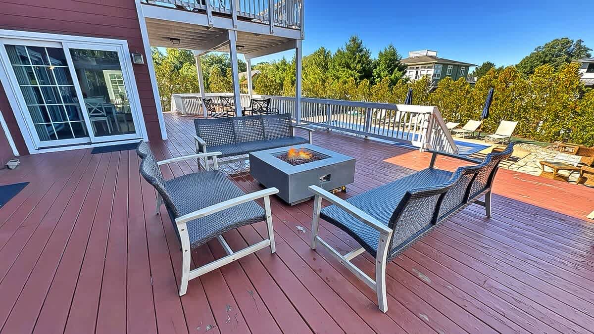 Expansive red-stained wooden deck featuring a modern fire pit surrounded by dark wicker outdoor seating, with lush green trees and a glimpse of a pool in the background under a clear blue sky.