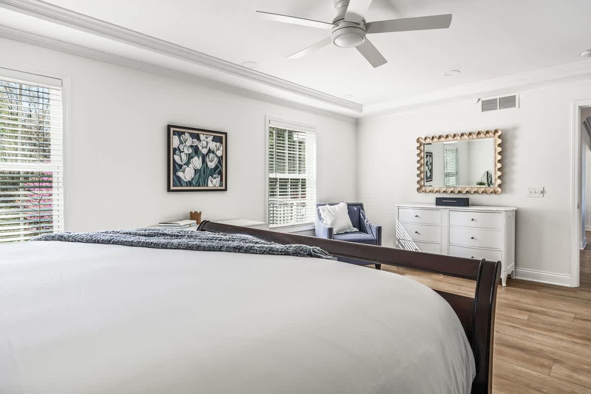 View of a bedroom with a dark wood bed, white bedding, and light wood floors, showing a white dresser with a decorative mirror and a blue armchair, with windows providing natural light.