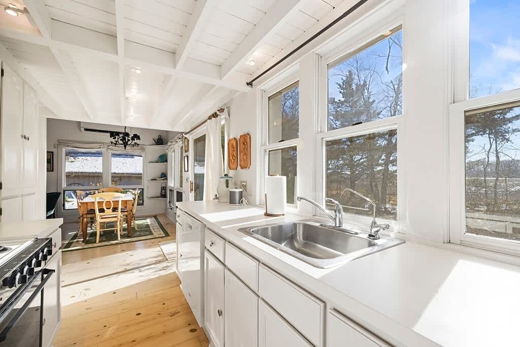 A bright kitchen with white cabinets and light wood floors features a stainless steel sink beneath large windows, offering a glimpse into the adjacent dining area with a wooden table.