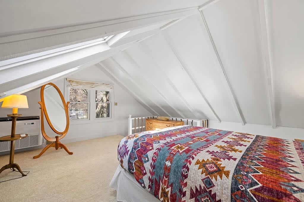 A bright attic bedroom features a sloped white paneled ceiling, a bed with a colorful patterned quilt, and a large oval mirror. A window offers a view of trees, and a small side table with a lamp is next to the bed.