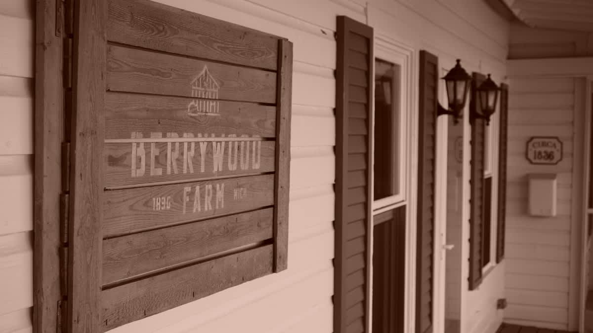 A wooden sign with "BERRYWOOD FARM" and "CIRCA 1836" is mounted on a white building with dark shutters and two wall-mounted lanterns. The image has a sepia tone.