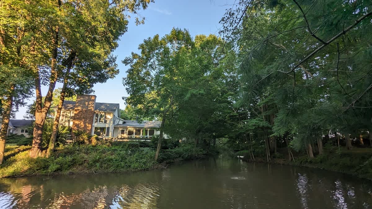 A serene river or canal flows through the property, flanked by lush green trees, with a large house featuring a brick chimney visible on the far bank under a clear blue sky.