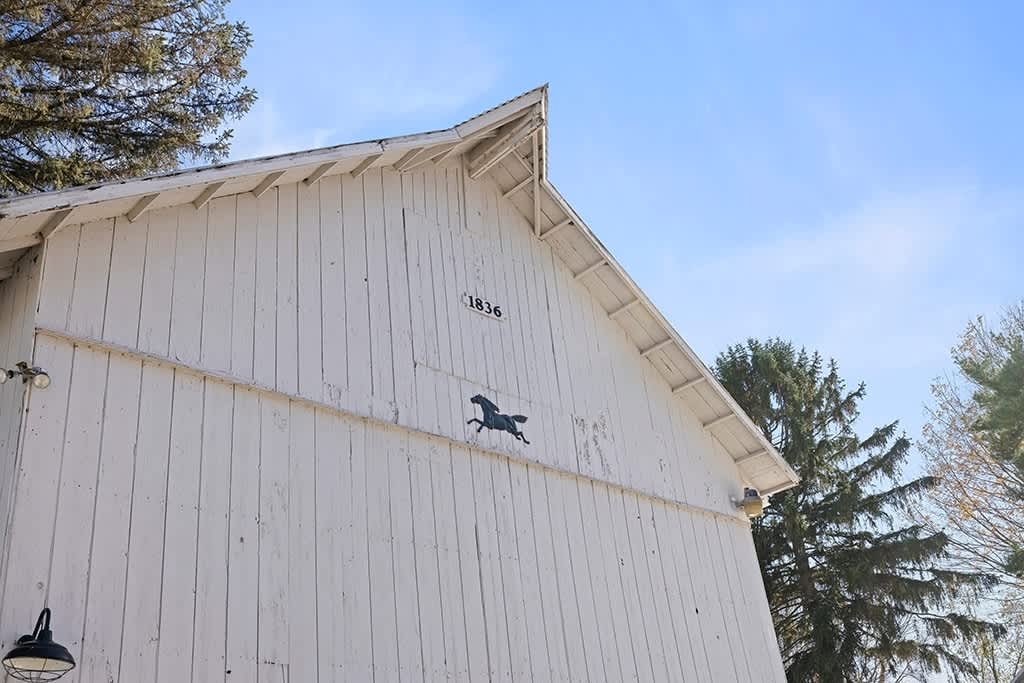 A close-up of the white barn's gable end, featuring vertical siding, a decorative horse silhouette, and the year "1836" inscribed near the peak. The bright blue sky and tree branches are visible above.