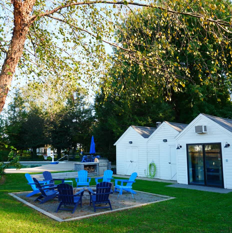 A cozy outdoor fire pit area features several blue Adirondack chairs arranged around a central fire pit, set on a gravel base near small white sheds and a swimming pool in the background.
