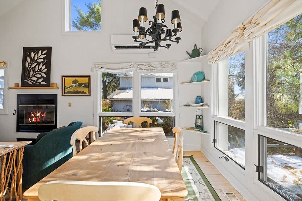 A bright dining area features a long wooden table and chairs, with large windows offering views of a snowy landscape, and decorative shelves on the wall. A black chandelier hangs from the vaulted ceiling.