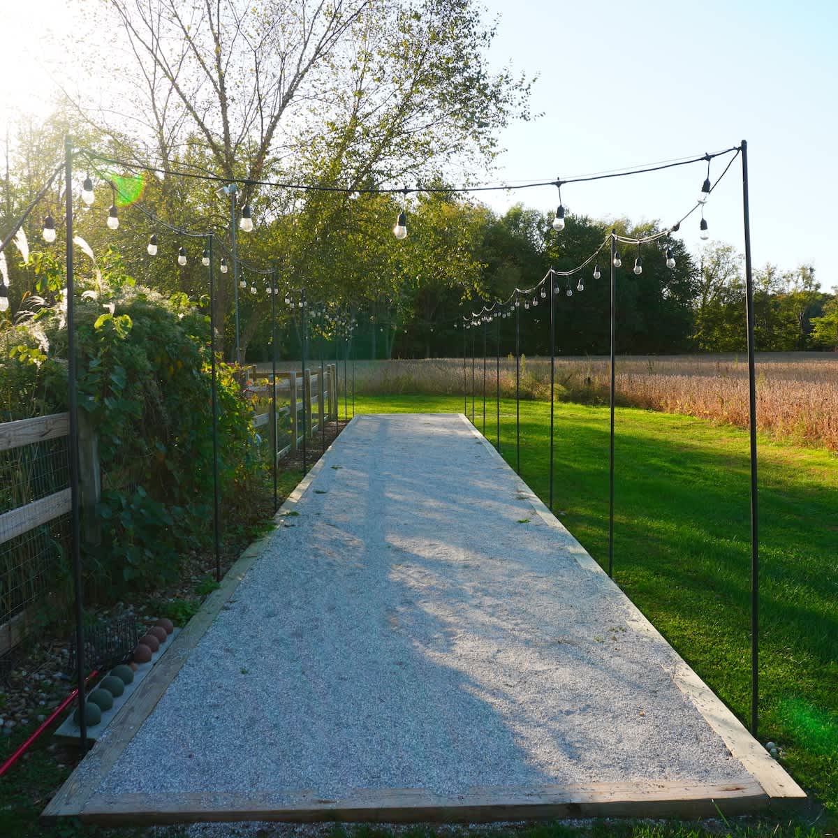 A long, gravel bocce ball court is illuminated by string lights strung between tall poles, set in a sunny grassy area adjacent to a cultivated field.
