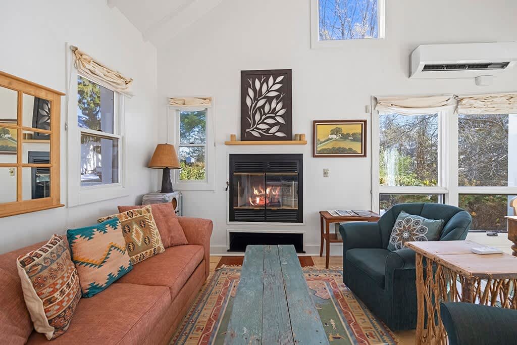 A cozy living room features a warm fireplace with a dark mantel, a rust-colored sofa with patterned pillows, a teal armchair, and large windows offering views of the outdoors.
