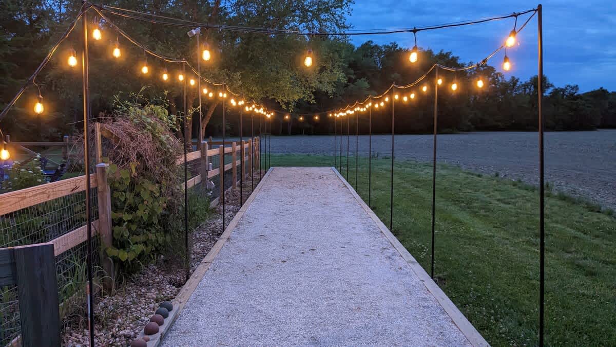 The gravel bocce ball court is warmly lit by glowing string lights at dusk, creating an inviting atmosphere in the grassy outdoor space next to a dark field.