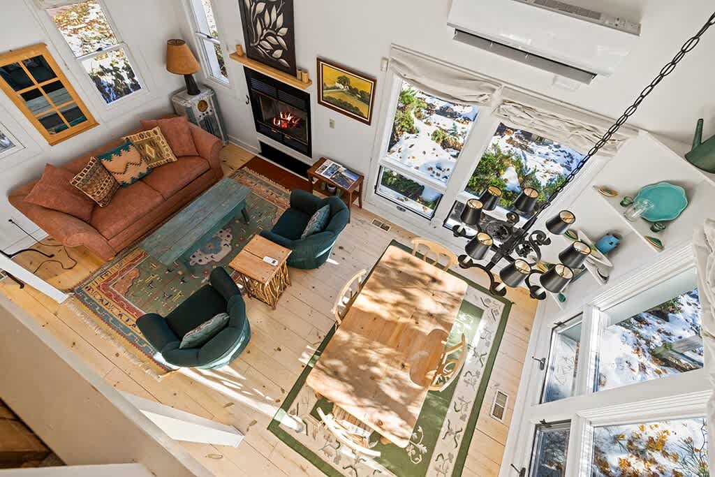 An overhead view captures the open-concept living and dining space, showcasing a large wooden dining table, a colorful area rug defining the living area with a sofa and armchairs, and a fireplace.