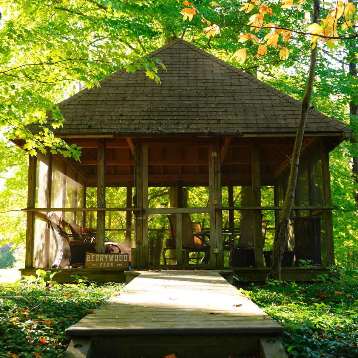 A rustic wooden screened gazebo with a shingled roof is nestled among vibrant green trees, featuring a wooden walkway leading to its entrance and a "BERRYWOOD FARM" sign inside.
