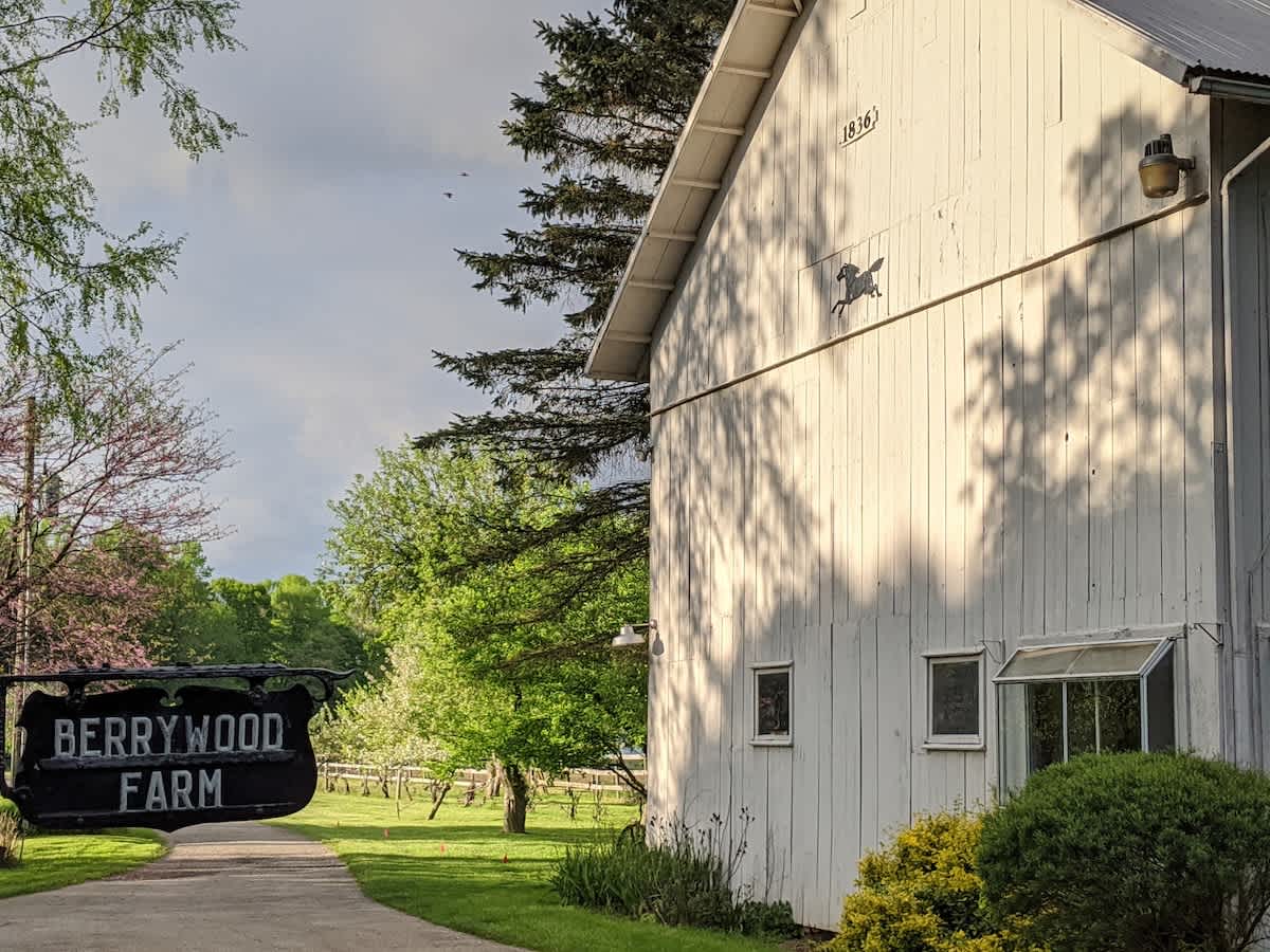 A white wooden barn, marked with "1836" and a horse silhouette, stands partially shaded by trees, with a prominent "BERRYWOOD FARM" sign in the foreground leading to a gravel path.
