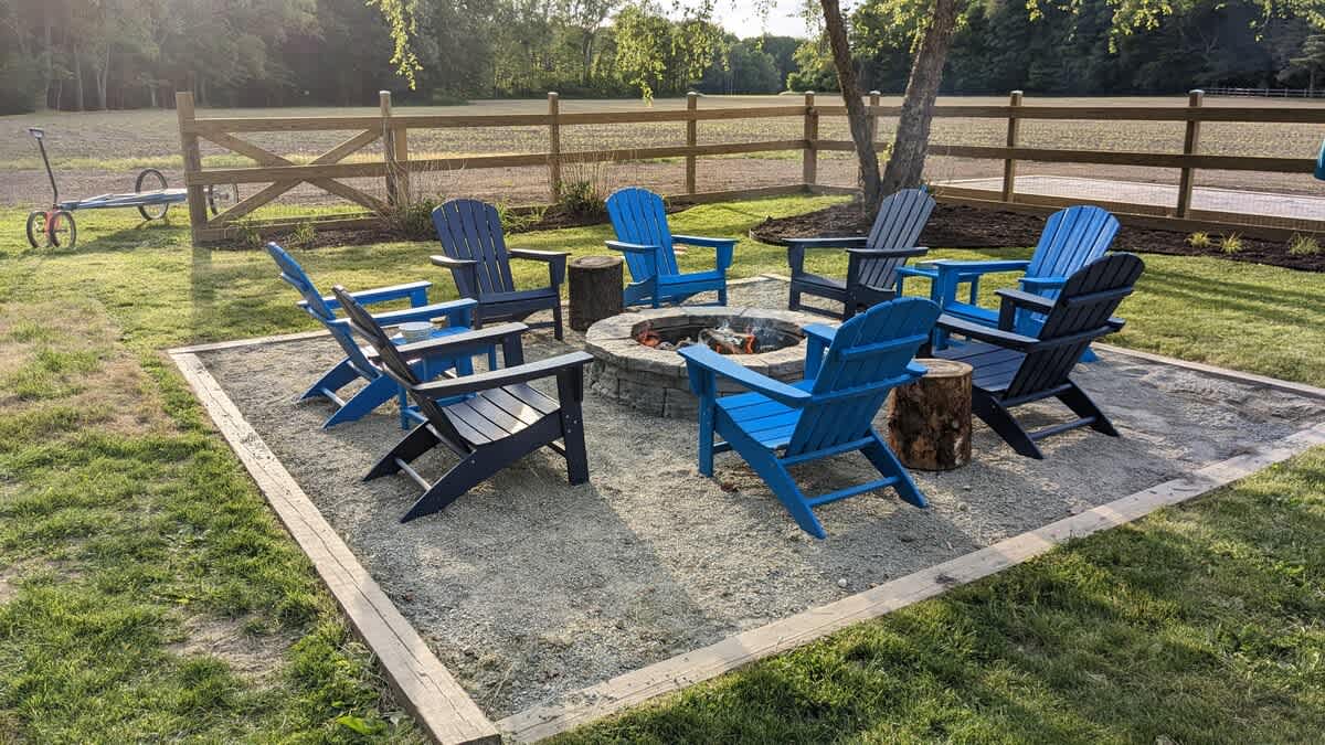 A circular stone fire pit is surrounded by eight blue and dark blue Adirondack chairs on a gravel patio, set within a grassy yard with a wooden fence in the background.