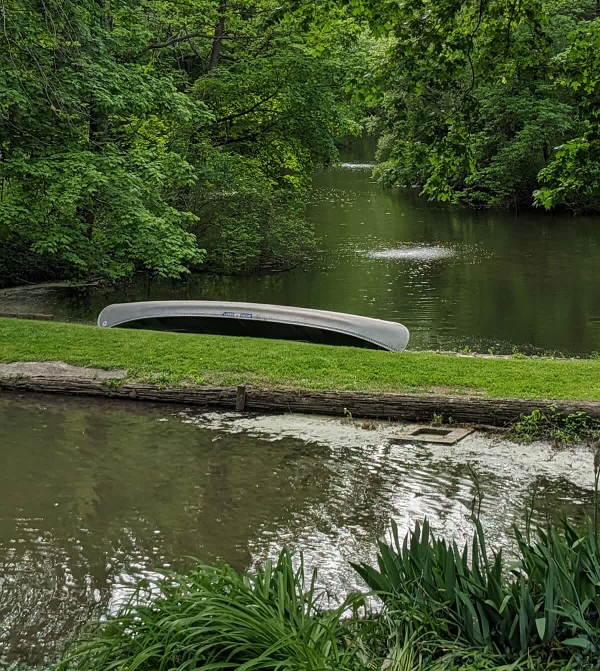 A light-colored canoe is stored upside down on a lush green grassy bank beside a calm, tree-lined river with a small fountain in the distance.