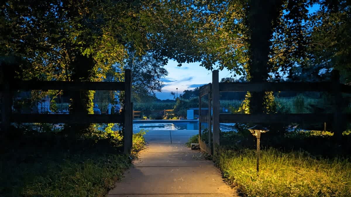A lighted pathway leads through a rustic wooden gate towards a swimming pool area, visible in the distance under a twilight sky, surrounded by dark trees.