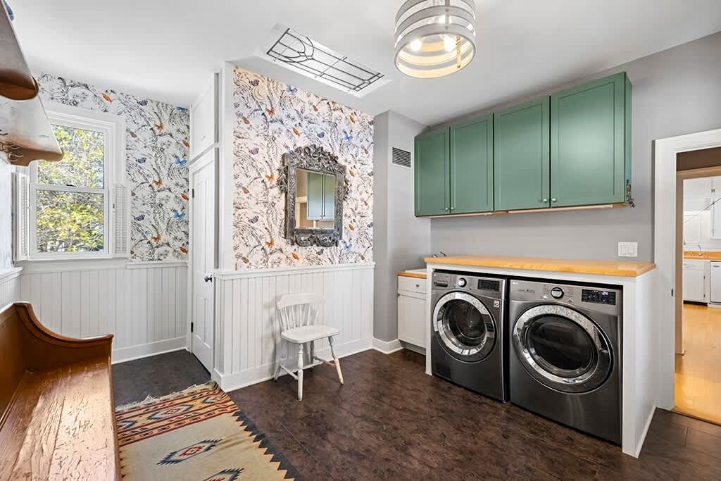 Bright laundry room with dark wood-look flooring, white wainscoting, and patterned wallpaper. It features a front-loading washer and dryer under a wooden countertop, green upper cabinets, and a small bench.
